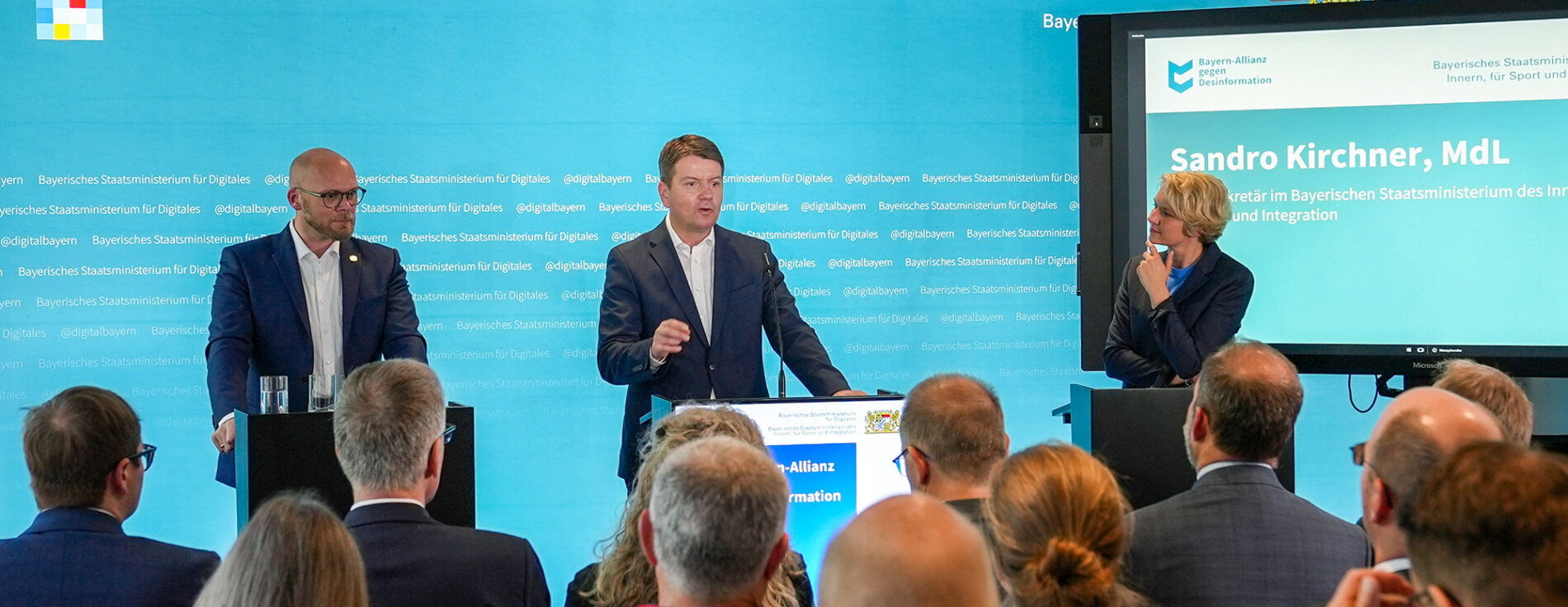 30. April 2025: Innenstaatssekretär Sandro Kirchner bei der Pressekonferenz zur Verstärkung der Bayern-Allianz gegen Desinformation: Gemeinsam mit Digitalminister Dr. Fabian Mehring stellt er neue zivilgesellschaftliche Partner vor, die mit ihrer breiten gesellschaftlichen Reichweite aktiv zur Aufklärung beitragen und damit ein starkes Zeichen für den Schutz unserer Demokratie setzen. Staatssekretär Sandro Kirchner (rechts), Staatsminister Dr. Mehring (links) stehen vor einer Pressewand mit Blick zum Publikum - rechts steht eine Moderatorin